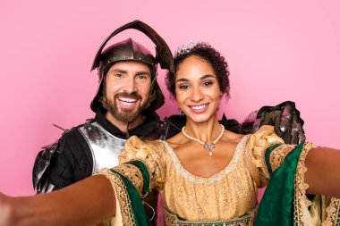 Young couple wearing royal and knight-inspired outfits take a fun selfie, depicting romantic bonding and amusement in a historical theme against a vibrant pink background. clipart