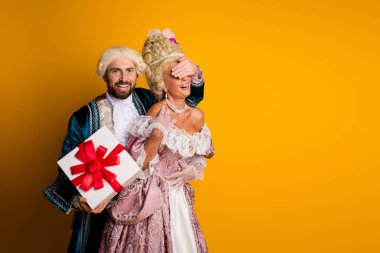 A young couple dressed in 18th-century noble attire sharing a joyful moment with a surprise gift exchange against a bright yellow background. clipart