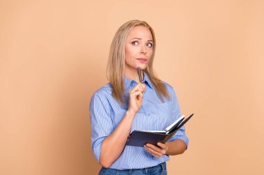 A stylish woman in a blue striped shirt holding a notebook and pen, thoughtfully planning or writing. Professional and creative setting with a warm beige-toned background. clipart