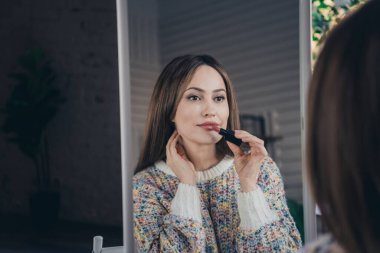 A young woman wearing a colorful sweater applies lipstick while looking into a mirror, enjoying a relaxed moment indoors. clipart