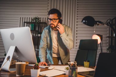 Focused businessman working on tasks, speaking on the phone while using a desktop computer in an organized office setting. clipart