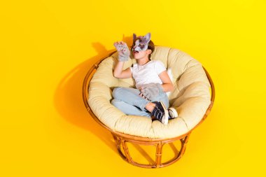 A youthful girl wearing a white shirt and jeans, with a playful cat mask, rests comfortably in a wicker papasan chair on a striking yellow background. clipart