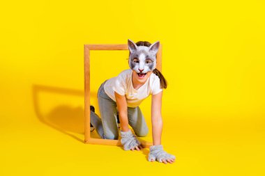 Playful young girl wearing a cat mask kneeling through a wooden frame on a bright yellow background, showcasing her creative and joyful spirit in a cosplay environment. clipart