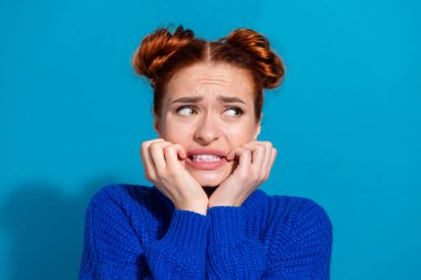 A freckled young woman with her hands near her mouth, dressed in a blue sweater, exudes an expression of nervous uncertainty against a vivid blue background. clipart