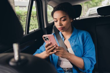 A young woman sitting in a car, using her smartphone. She looks focused and relaxed, enjoying her urban travel experience on a sunny day. clipart