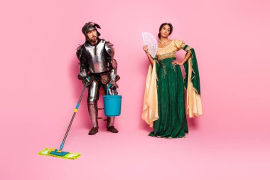 Amusing image of a woman in elegant medieval dress next to a man wearing knight armor cleaning with modern tools, against a vibrant pink background. clipart