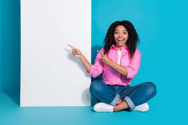 A young woman with curly hair in casual attire points excitedly at a white board against a vibrant blue background, exuding happiness and positivity. clipart