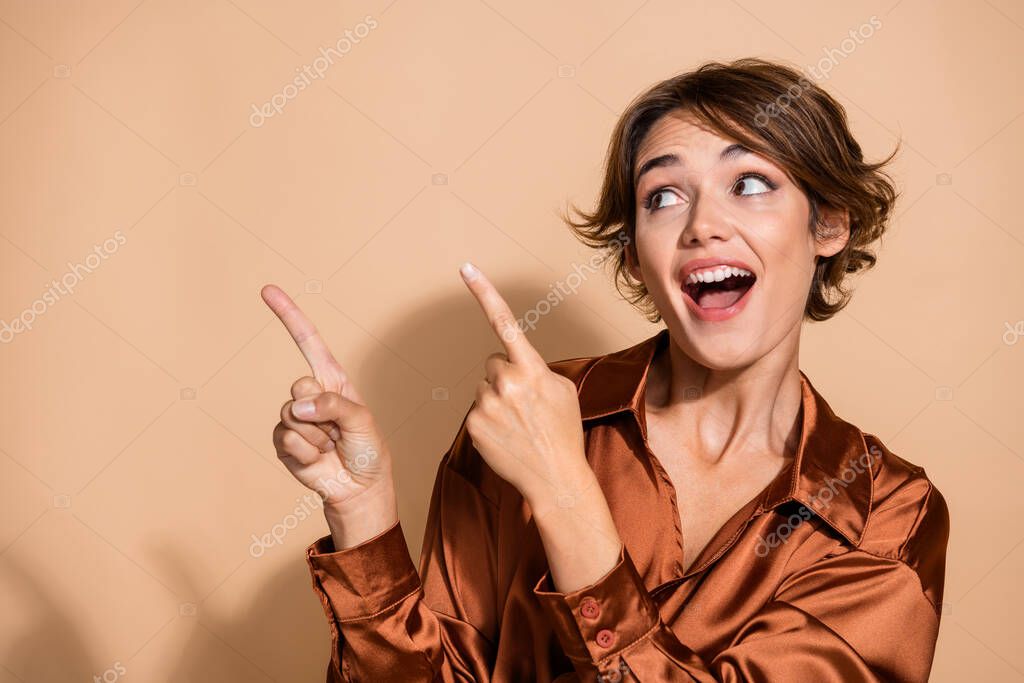 Photo of impressed funky woman wear silky blouse pointing two fingers looking empty space isolated beige color background.