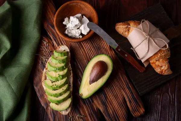 Croissant with avocado, sesame and flax seeds on wood background. Cereal croissant, plate of cream cheese and knife