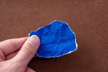 Hand holding blank blue paper on brown background. Top view, copy space.