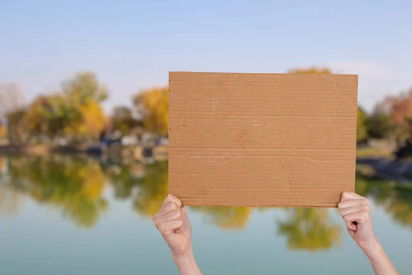 Hand holding empty cardboard sign against blurry lake background