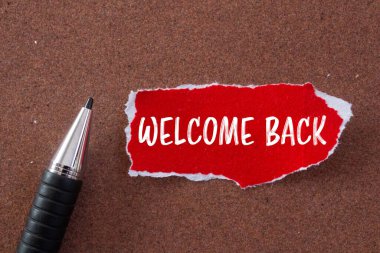 Welcome back message written on ripped torn red paper piece and a pen with brown background. Conceptual welcome back symbol. Copy space. clipart