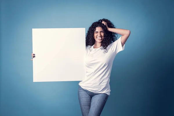 Beautiful curly woman holding blank billboard isolated on blue background making the tongue in a funny way