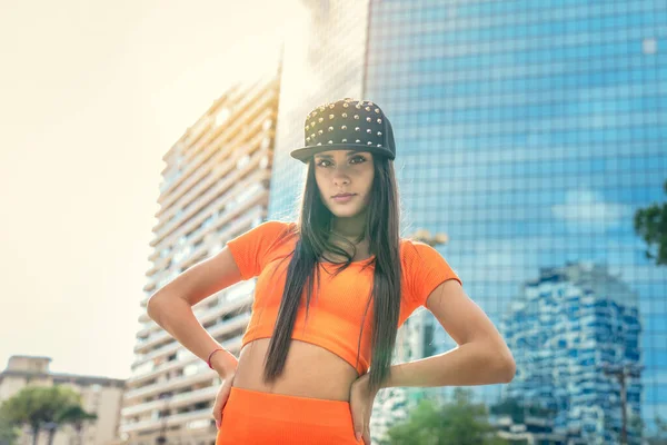 照片拍摄时尚的高加索嘻哈舞者站在橙色的腿和顶部体操套衫和黑色的帽子与城市为背景 城市建筑物射击健身模型 — 图库照片