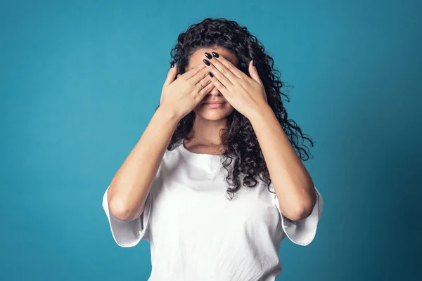 Shot of serious confident curly woman, wears casual basic solid white t-shirt, covering her face isolated over blue background. Expressions