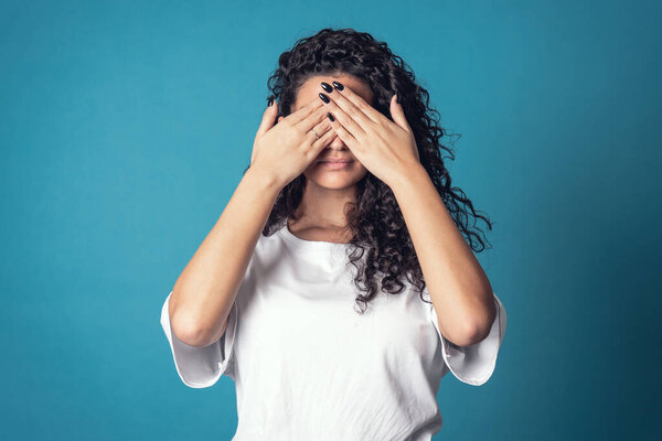 Shot of serious confident curly woman, wears casual basic solid white t-shirt, covering her face isolated over blue background. Expressions