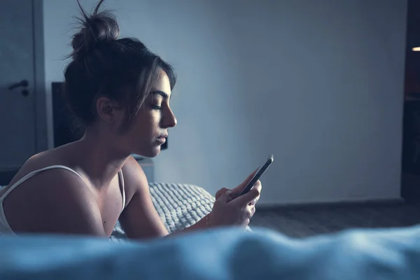 Caucasian girl passing the night messaging in her bedroom watching the cellphone device. A serious girl alone on her bed chatting holding her smart phone in the dark of her room with a soft light