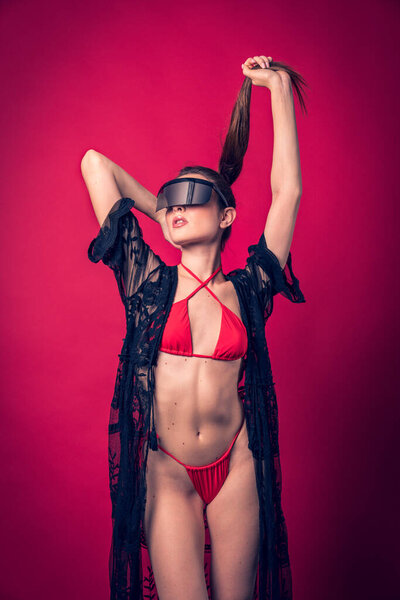 Studio photo shoot of beautiful brown hair model posing wearing red swimwear and futuristic glasses with red background behind