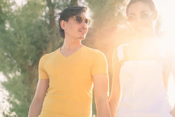 ハンサムな男は 休暇中の晴れた日に公園でサングラスをかけて手を握って美しい女性と歩いています 若い大人のカップル一緒に緑の木と太陽のフレアに囲まれて歩く — ストック写真