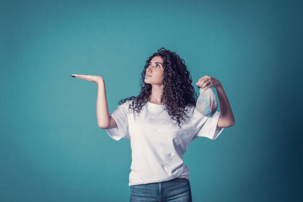 Студия снимка красивой уверенно кавказской кудрявой модели женщины носит синие джинсы и повседневную простую белую футболку, изолированную на синем фоне. Красивая брюнетка держит в руках маленькую планету.