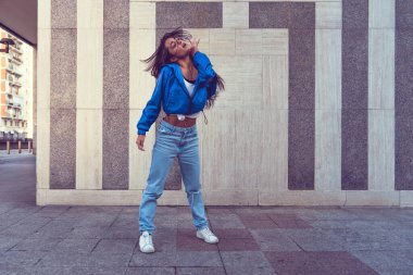 Modaya uygun beyaz hip hop dansçısının mavi ceket, kot pantolon, siyah bluz ve beyaz tişört içinde eğlenirken çekilmiş bir fotoğraf, arka plan olarak kentin tadını çıkar. Şehir binaları hareket eden fitness modelini çekiyor