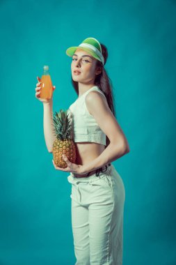 Stüdyoda siyah mayo, beyaz pantolon ve ananas ve meyve suyu tutan yelek giymiş güzel kahverengi saç modelinin moda fotoğraf çekimi. Stüdyo ışığı bir fotoğraf stüdyosunda güneşi canlandırıyor..