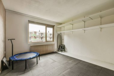 Trambolin ve egzersiz bisikletiyle dolu geniş bir odada pencerenin yanındaki duvar rafı.