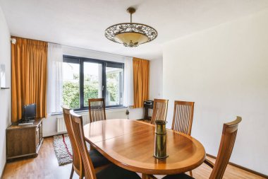 Modern oturma odasında pencerenin yanında kanepesi olan siyah kolye lambası altında çiçek vazosu olan ahşap bir masa.