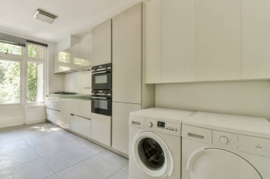 Gündüz vakti çamaşır makinesinin yanında modern dolaplar ve aletlerle aydınlık mutfağın içinde.