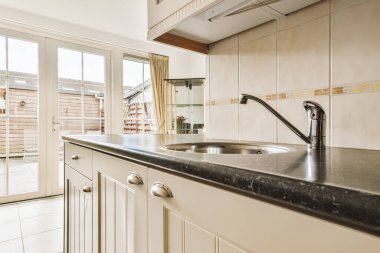 Resmin ortasında beyaz dolapları ve siyah tezgahları olan bir mutfak verandaya açılan açık bir kapıdır.