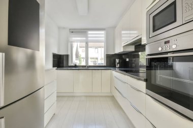 Duvarında beyaz dolapları ve siyah tezgahları olan modern bir mutfak. Odadaki paslanmaz aletlerle birlikte.