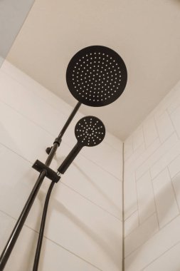 Duş başlığı ve termostaer. Arkasında beyaz fayanslar olan bir banyoda siyah bir el var.