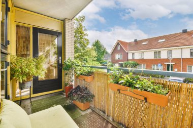 Arka planda mavi bir gökyüzüne karşı tırabzanlar ve saksılarla dolu bir balkon dışarıdan çekilmiştir.