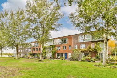 Bir apartman kompleksi. Önünde ağaçlar, yerde çimenler, mavi gökyüzünün altında yemyeşil çimlerle çevrili.