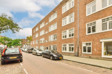 Beyaz süslemeli ve pencereli kırmızı tuğlalı bir binanın önüne park etmiş birkaç araba var.