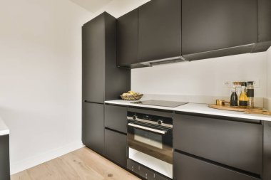 Modern bir mutfak, siyah dolaplar ve duvarında beyaz tezgahlar, odanın ortasında bir fırın var.