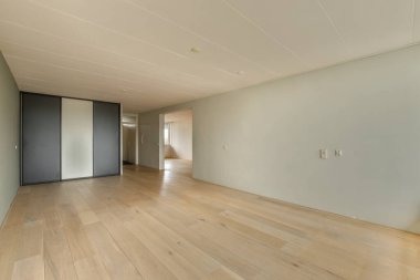 Ahşap döşemesi ve duvarlarında beyaz boya olan boş bir oda. Köşesinde siyah bir kapı var.