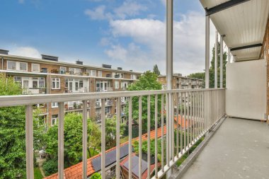 Arka planda ağaçlar ve evler olan bir balkon, aşağıdaki sokağa bakan bir apartman penceresinden alınmış.