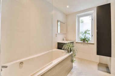 Küvetli bir banyo ve penceresinde bitkiler var. Küvetin yanındaki duvara yapıştırılmış.