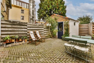 Güneşli bir günde arka bahçesinde sandalyeler, masalar ve ağaçlar olan bir teras var. Mavi bir gökyüzü var.