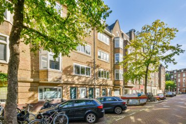 Bir apartmanın önündeki kaldırımlara ve ağaçlara park etmiş arabaların olduğu bir şehir caddesinde birinin üzerine yaslanmış bir bisikleti var.