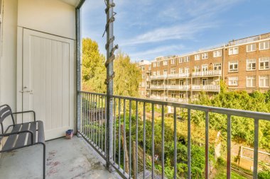 Ön planda sandalyeler ve ağaçlarla dolu bir balkon. Güneşli bir günde bir apartmanın penceresinden çekilmiş.