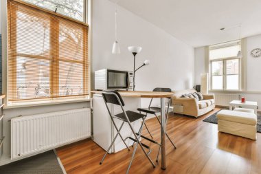 Pencerenin önünde küçük bir yemek masası da dahil olmak üzere ahşap döşemeli ve beyaz duvarlı bir oturma odası.