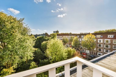 Arka planda ağaçlar ve evler olan bir balkon, aşağıdaki sokağa bakan bir apartman penceresinden alınmış.