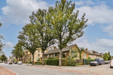 Fotoğrafın ortasında evleri ve ağaçları olan bir cadde sokağın karşısında park edilmiş arabalar var.