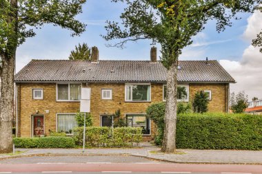 Önünde ağaçlar ve her iki tarafında da çalılar olan eski bir tuğla ev sokak perspektifinden alınmış.