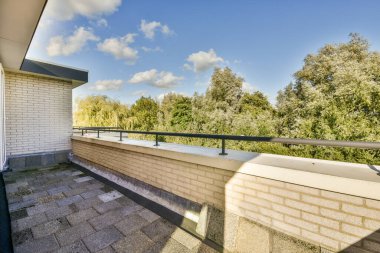 Arka planda ağaçlar ve yukarıda mavi gökyüzü olan bir balkon güneşli bir günde açık teras alanından görüldüğü gibi.