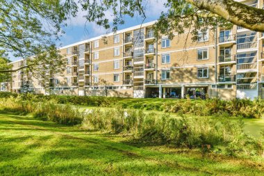 Güneşli bir yaz gününde, ön planda yeşil çimenler ve ağaçlarla dolu bir apartman kompleksi.