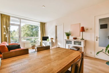 Oturma odası, koltuklar, sandalyeler ve büyük bir pencerenin önünde sokağa bakan bir masa.