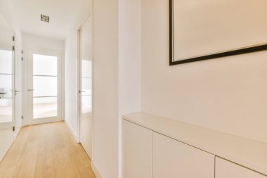 Beyaz duvarları ve kapının yanında ahşap döşemeleri olan boş bir koridor siyah çerçeveli bir ayna.